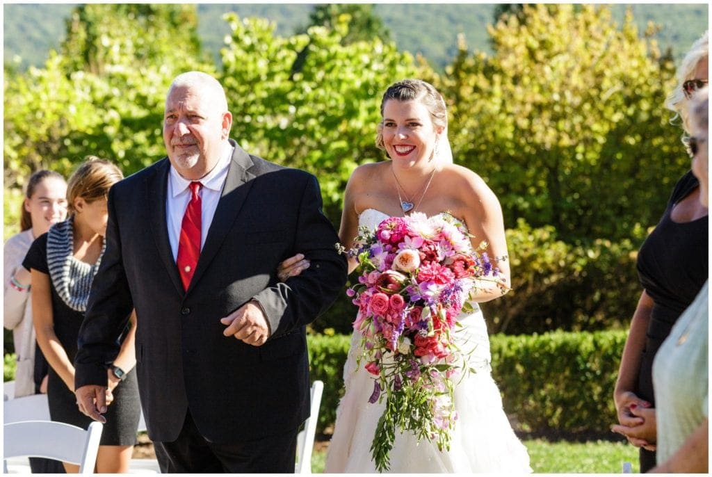 Bride walking down the aisle with gorgeous cascading pink and purple bouquet by Splints and Daisies.