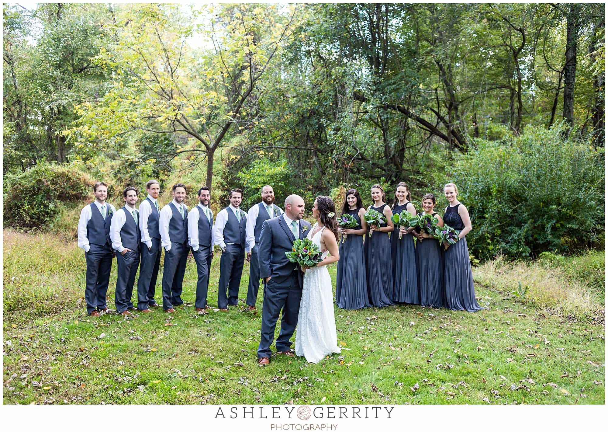 Bride & groom with their wedding party, groomsmen dressed in blueish grey vests & pants, bridemaids in deep periwinkle full length gowns, carrying vegetable bouquets