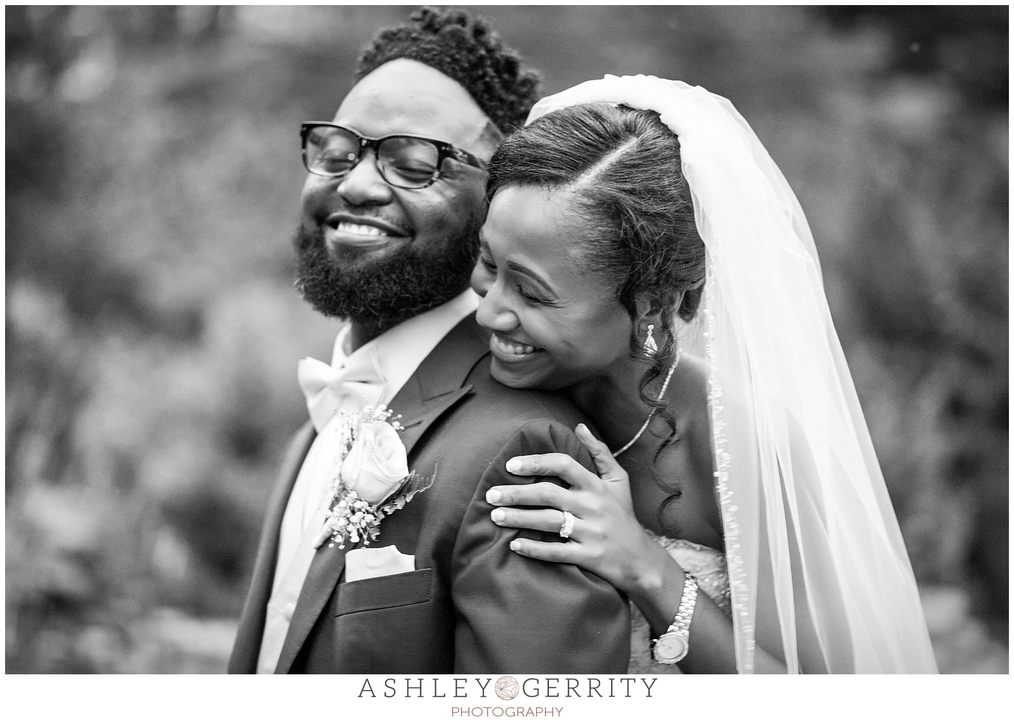 Black & white photo of bride hugging groom from behind and resting her head on his shoulder while smiling at the Philadelphia Museum of Art Azalea Garden