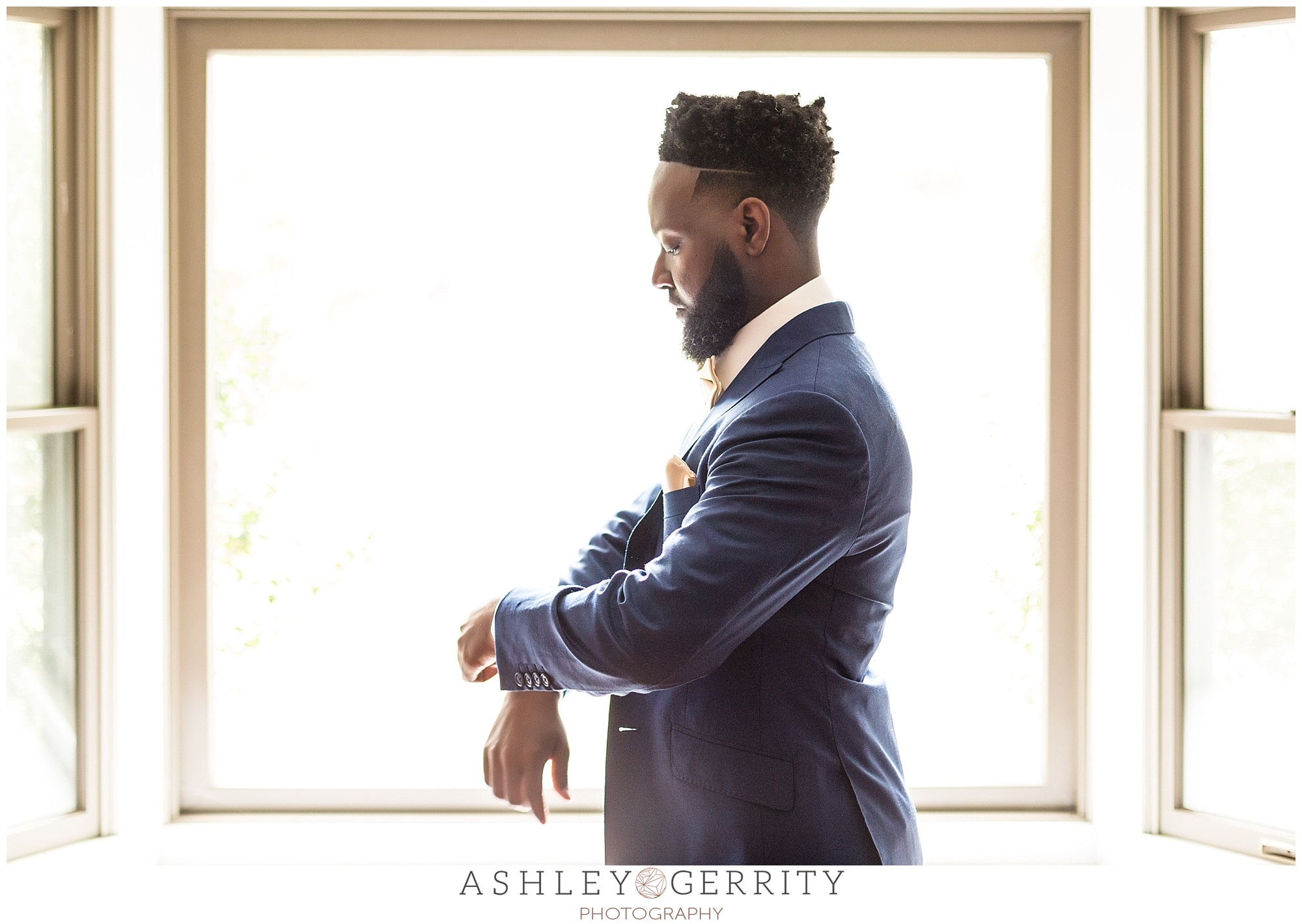 groom adjusts the sleeve of his wedding suit in front of a large picture window
