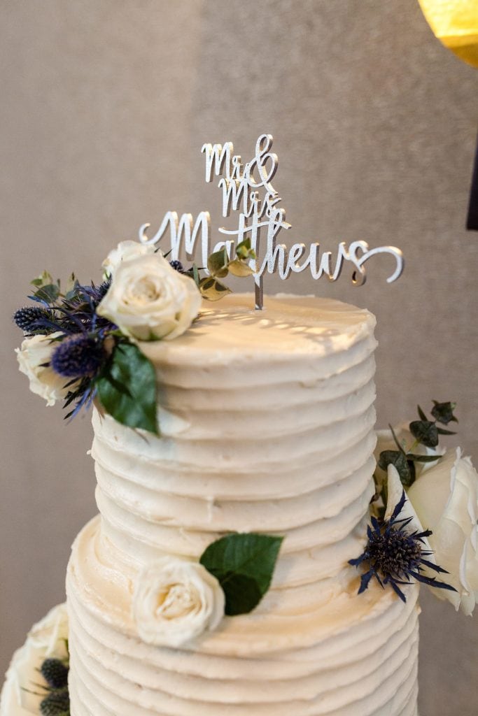 Tiered wedding cake covered in ridged vanilla buttercream with laser cut script cake topper and decorated with roses and thistle