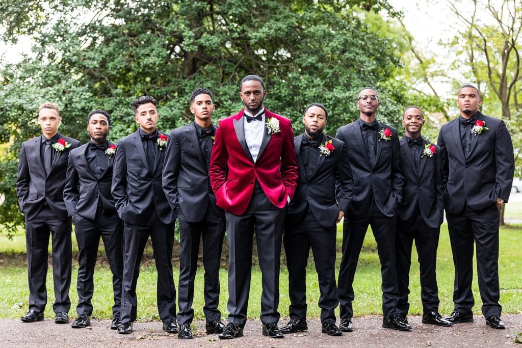 Groom in a red crushed velvet jacket with black lapels poses with his party of groomsmen all in black