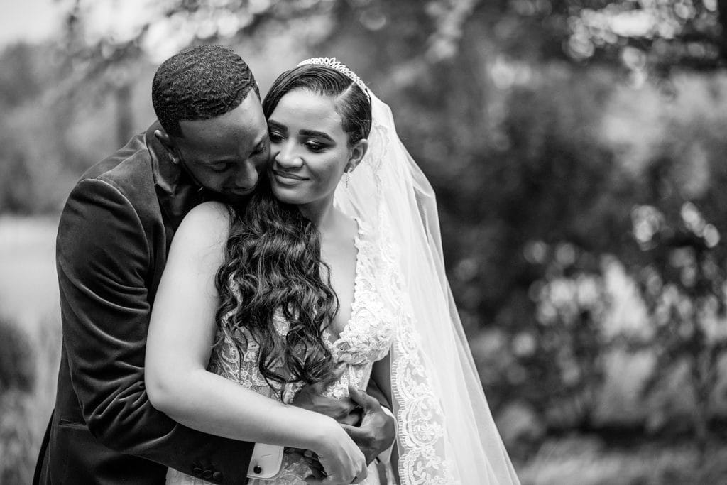 Groom embraces bride from behind and kisses her shoulder. Photographed in black & white.