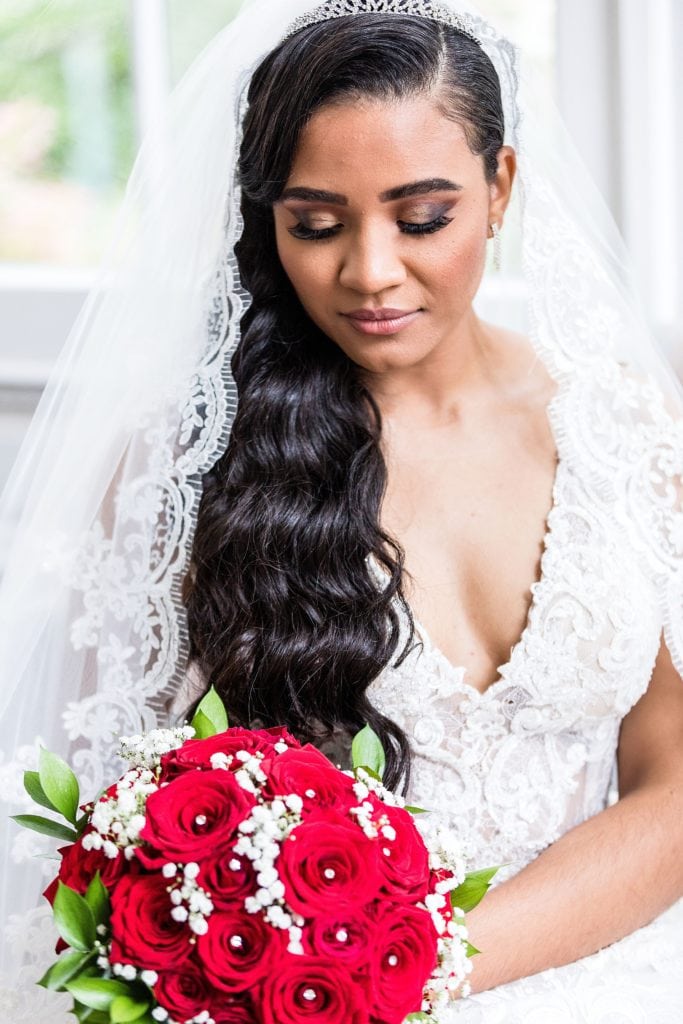Window lit portrait of a bride in a lace Galina Signature wedding dress while holding a bouquet of red roses