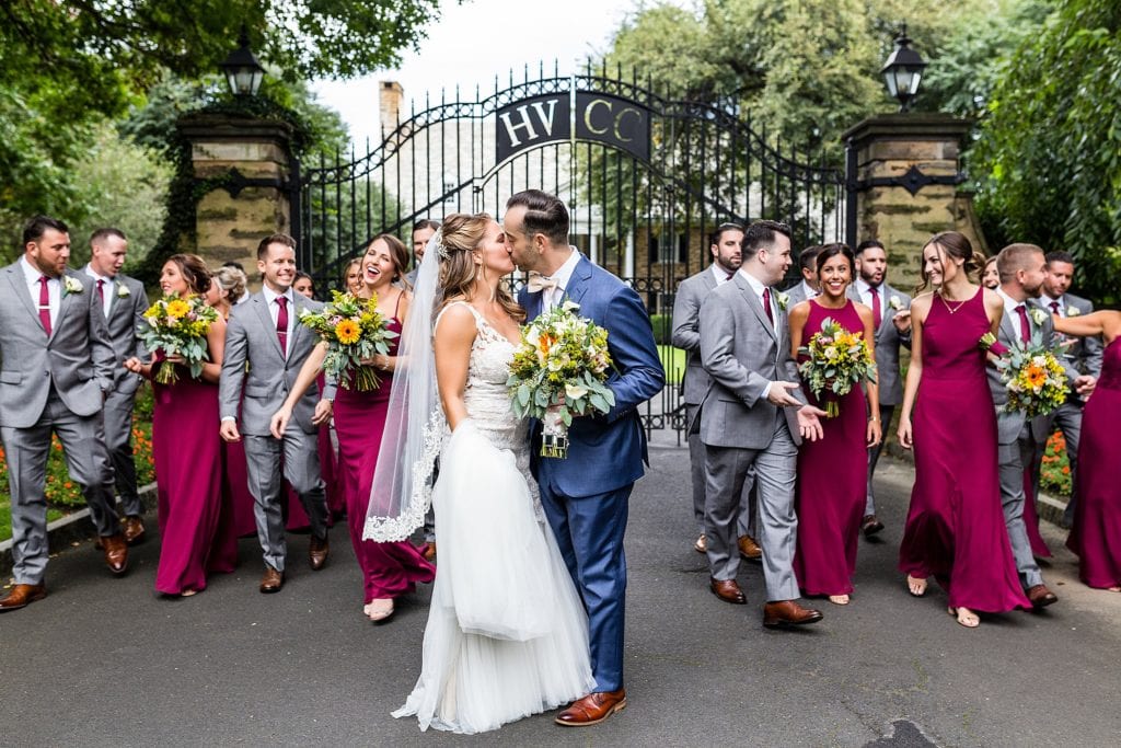 Bride & groom kiss and walk with their wedding party posing in front of the house and the gate at Huntingdon Valley Country Club