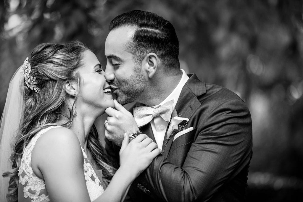 Bride & groom laugh as they kiss each other. Portrait photographed in black & white