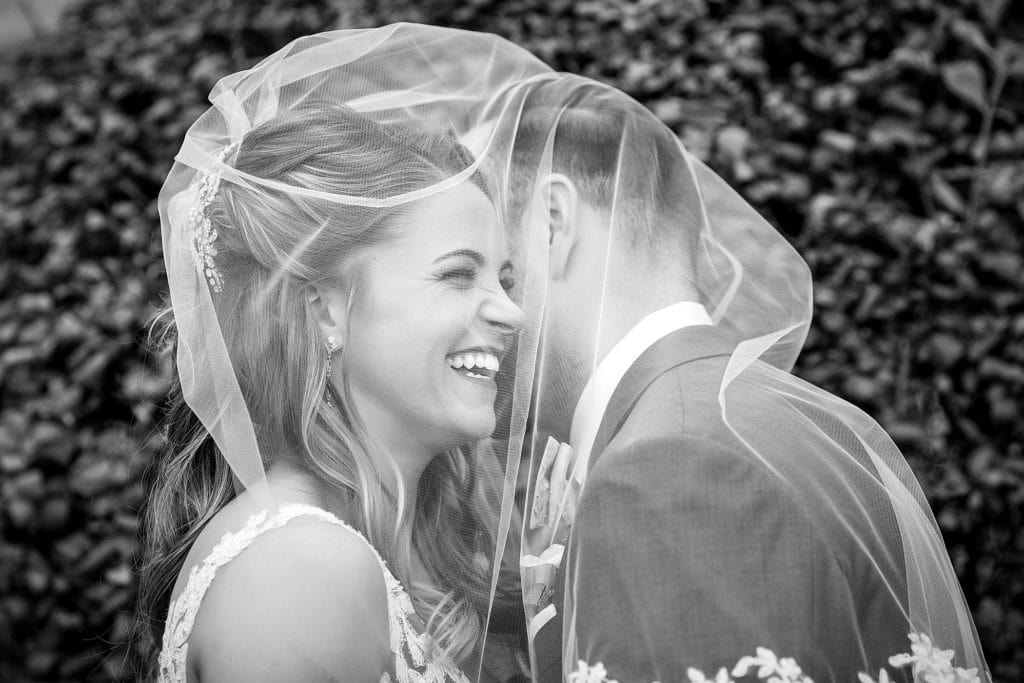 Bride & groom laugh together under the veil in front of a wall of greenery. Photographed in Black & white.