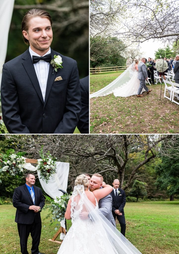 Groom smiles as a bride is walked down the aisle by her brother while her cathedral length lace veil trails behind her during her outdoor John James Audubon Center wedding ceremony