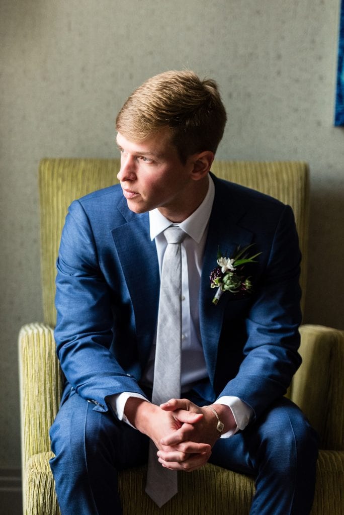 Portrait of a groom in a corduroy chair with a Succulent boutonnierre by Devon & Pinket against a blue tailored groom's suit and burgundy polka dotted handkerchief