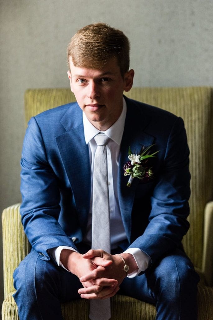 Portrait of a groom in a corduroy chair with a Succulent boutonnierre by Devon & Pinket against a blue tailored groom's suit and burgundy polka dotted handkerchief