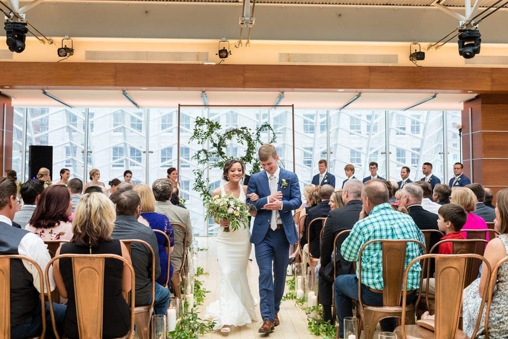 Bride & groom walk out together from the industrial ceremony space for Kimmel Center wedding showing a ceremony backdrop of copper pipe and greenery wreaths