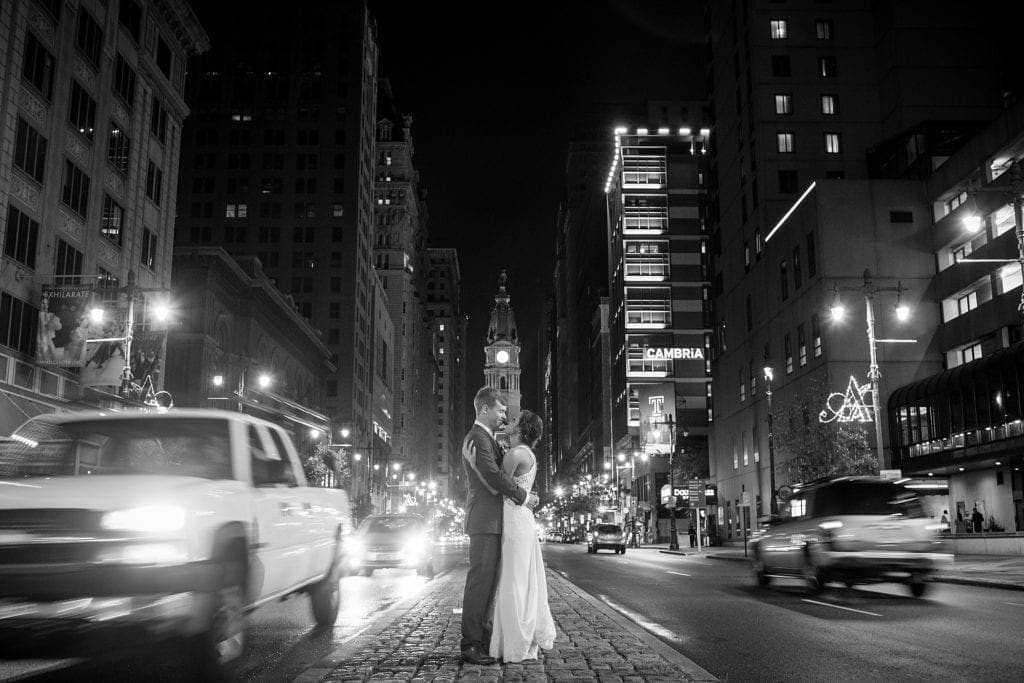 Black & white wedding photo of a bride & groom on Broad St in Philadelphia in front of City Hall as traffic rushes by