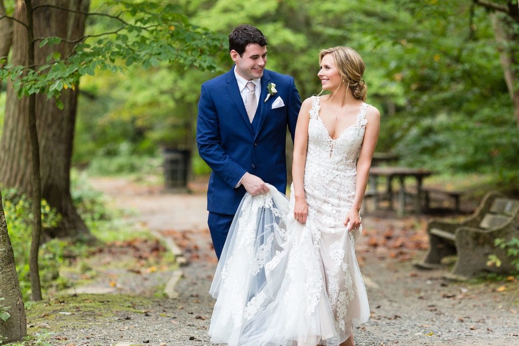 Groom carries the train of a bride's lace wedding dress for their Morris Arboretum wedding