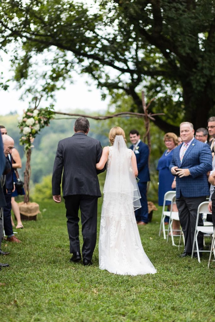 Groom smiles as bride walks down the aisle with her father during their Morris Arboretum wedding ceremony