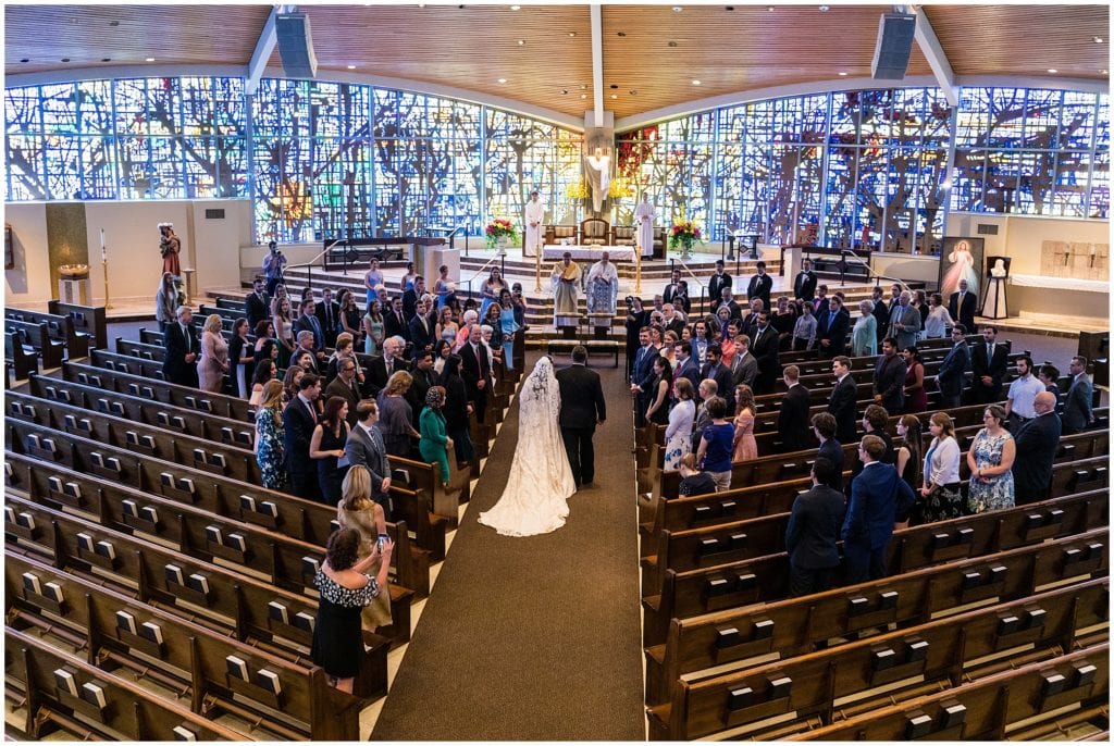 Wide angle photograph of bride and her father walking down the aisle in church wedding ceremony with huge stained glass windows