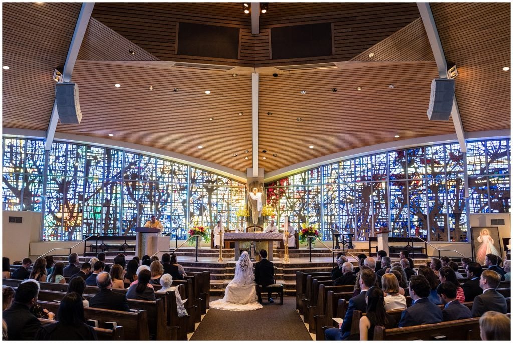 Wide angle photograph of bride and groom in church during wedding ceremony with huge stained glass windows