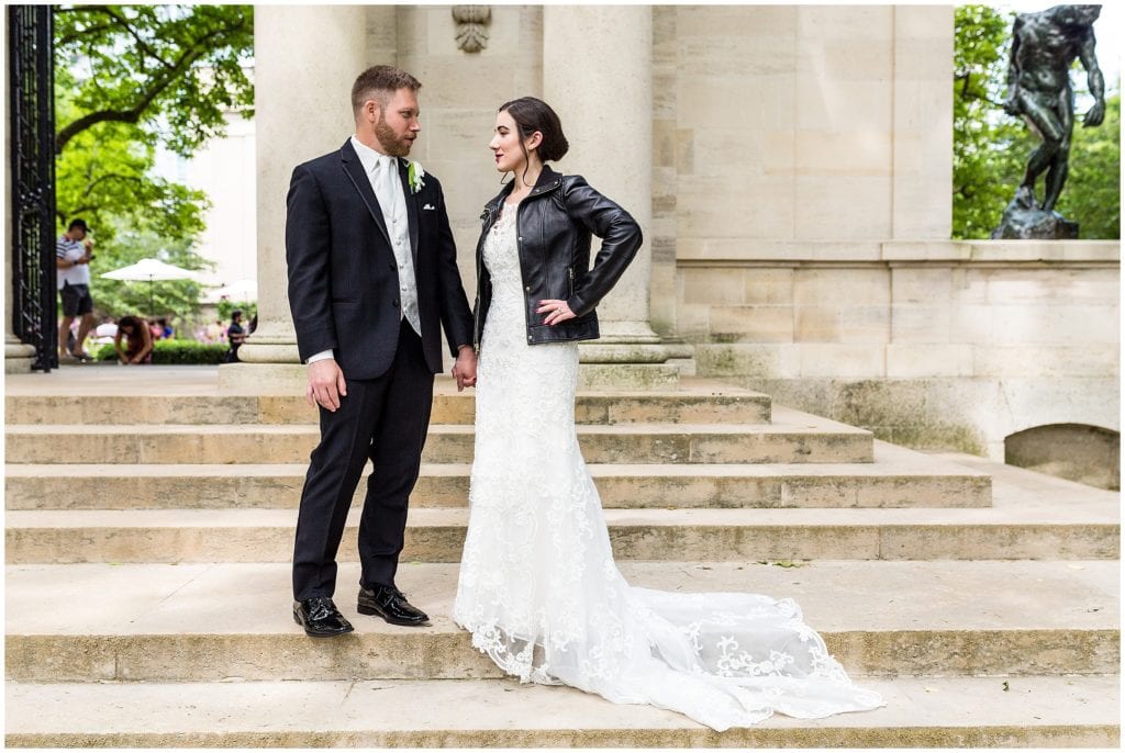 Bride rocking a leather jacket over her Maggie Sottero wedding dress while holding groom's hand on the steps of the Rodin Museum