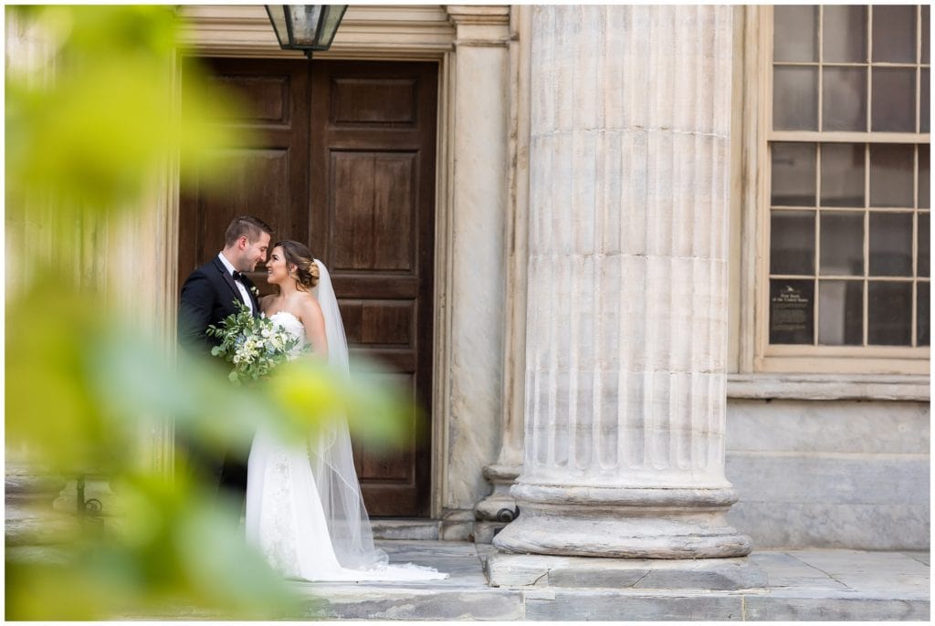 romantic outdoor portrait of bride and groom in front of marble pillars taken with greenery in the foreground