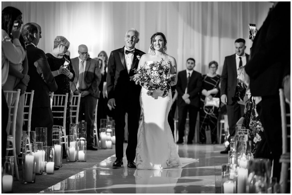 Black and white portrait of the bride and her father walking down the aisle for her wedding ceremony at the Lucy by Cescaphe