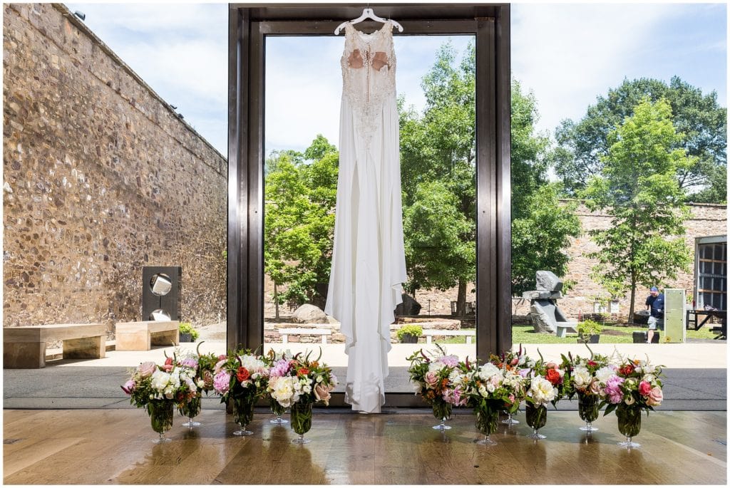 Bride's dress hanging in giant window at Michener Museum with Garnish by Catering by Design floral arrangements on the floor