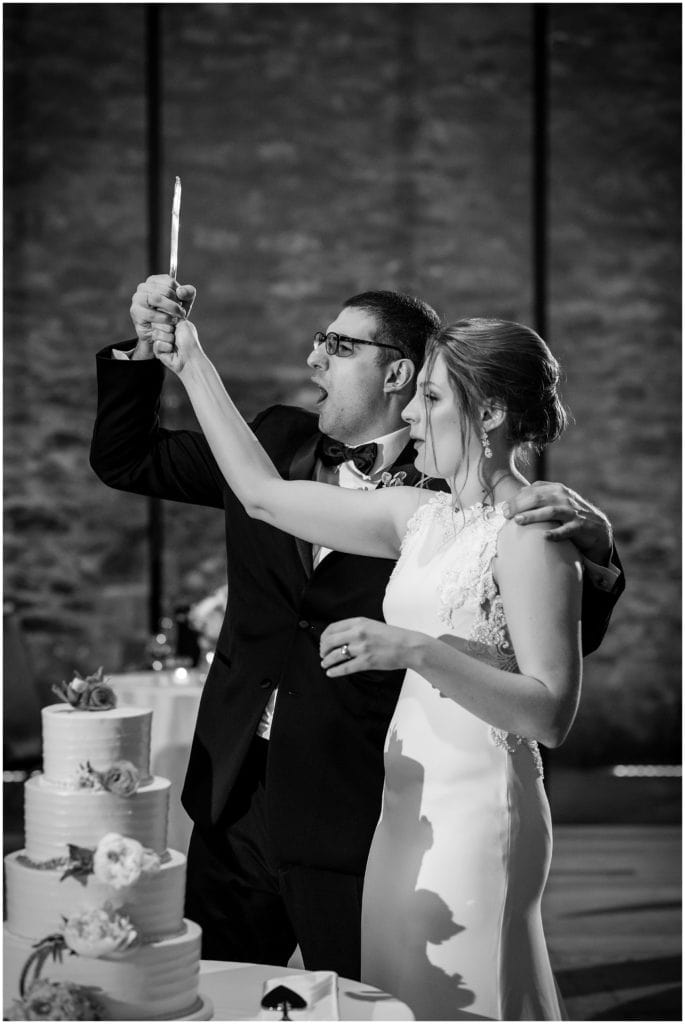 black and white image of groom playfully holding cake cutting knife aloft with his bride as they cut the cake
