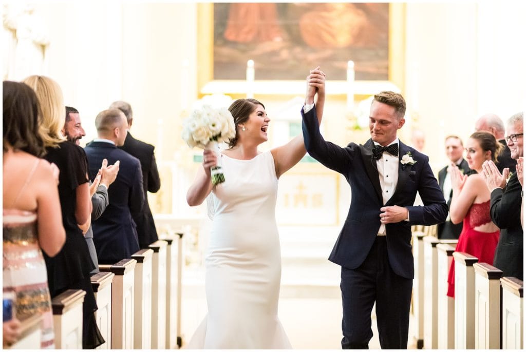 Bride and groom holding hands and cheering as the walk up the aisle after Catholic Church wedding ceremony