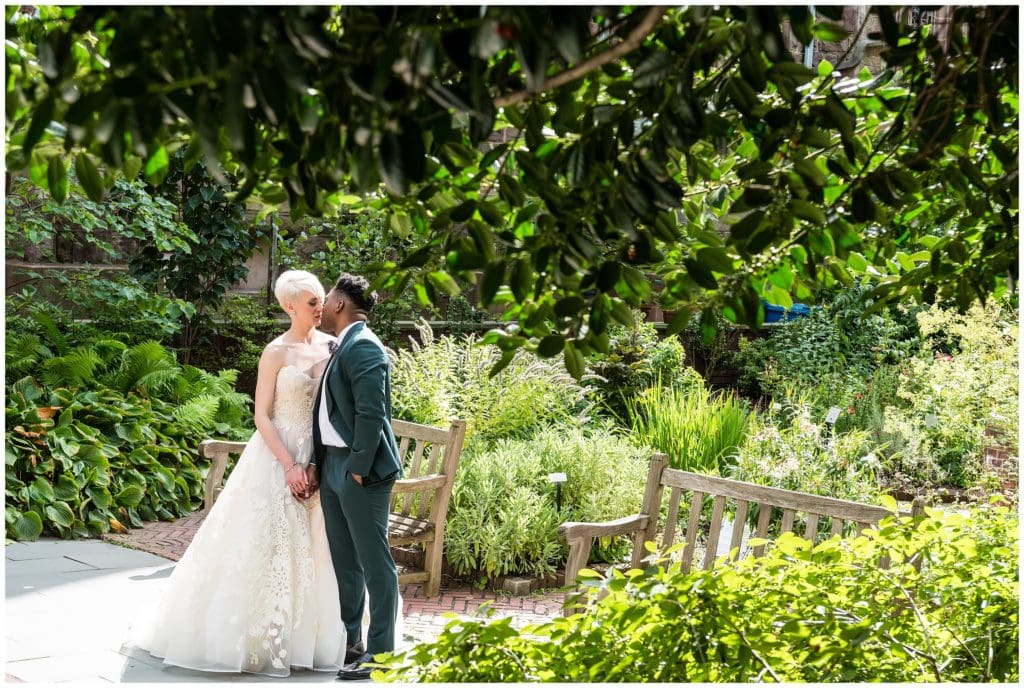 Bride and groom kissing outside through trees in urban green garden space