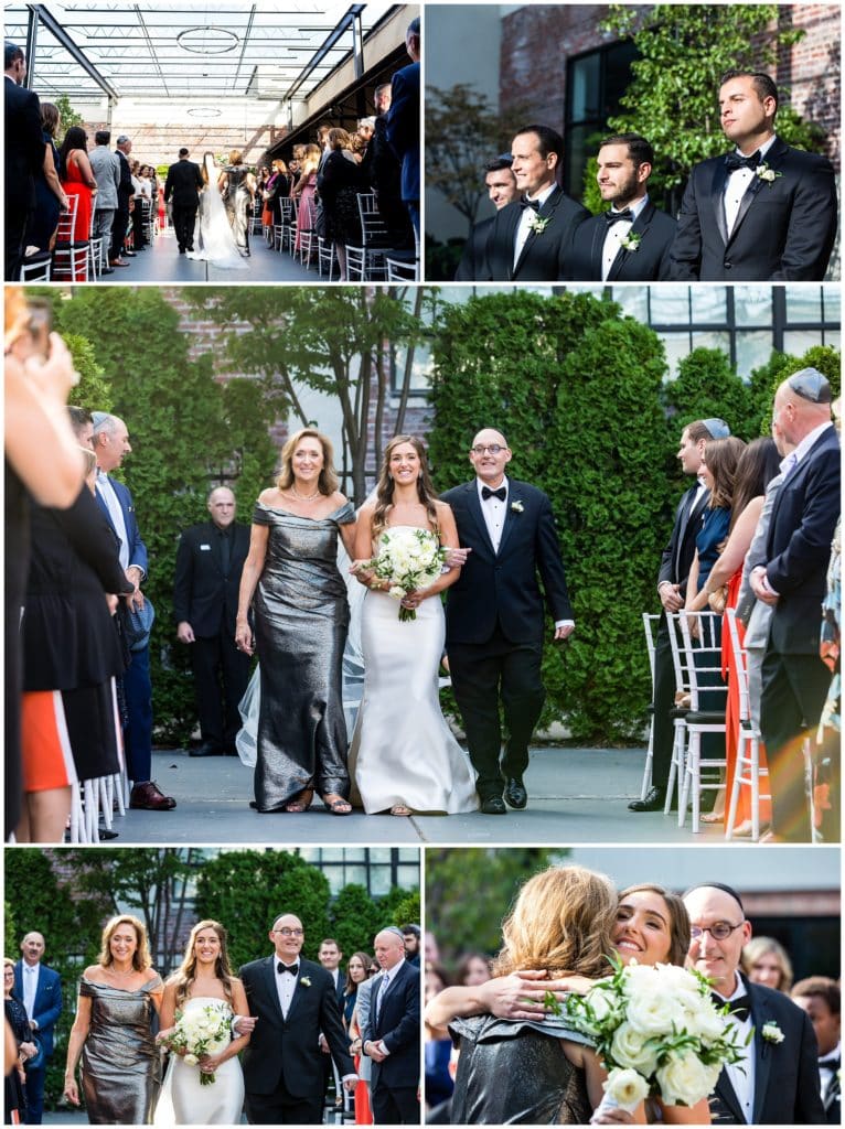 Bride being walked down the aisle by both her parents in outdoor courtyard Vie Jewish wedding ceremony