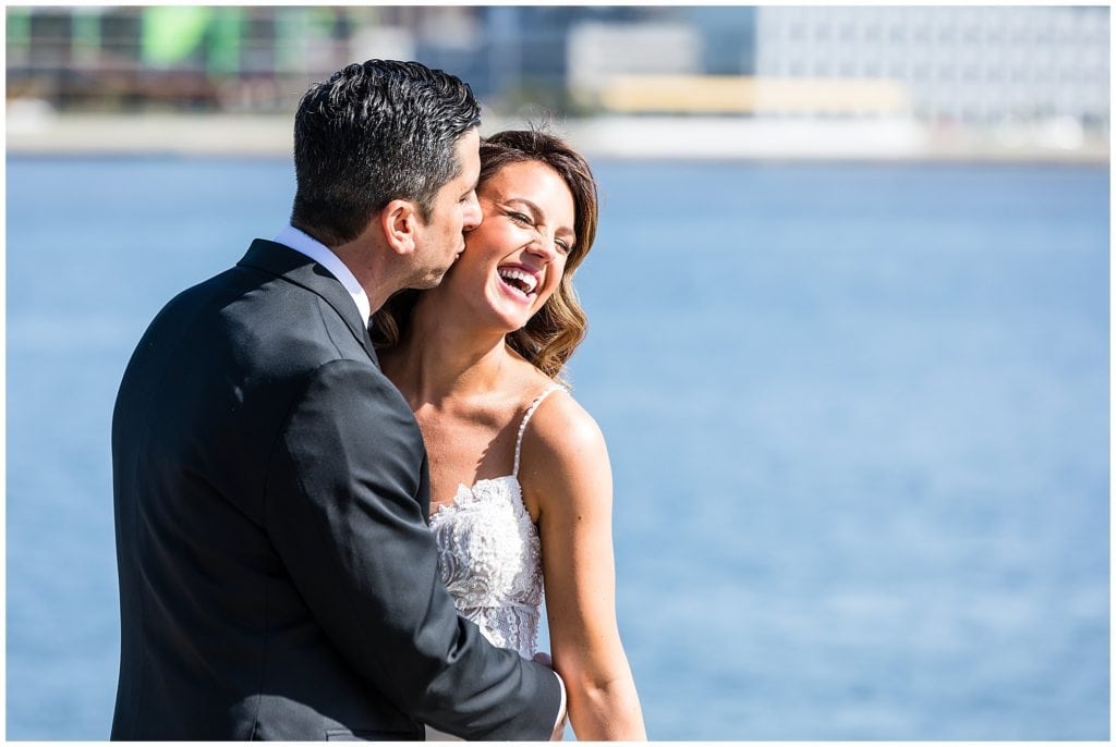 Bride laughing while groom kisses her on the cheek in front of river in Philadelphia