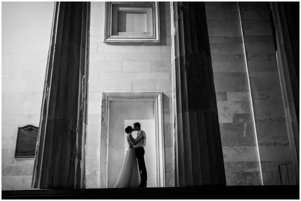 Dramatic black and white night portrait with bride and groom kissing between pillars