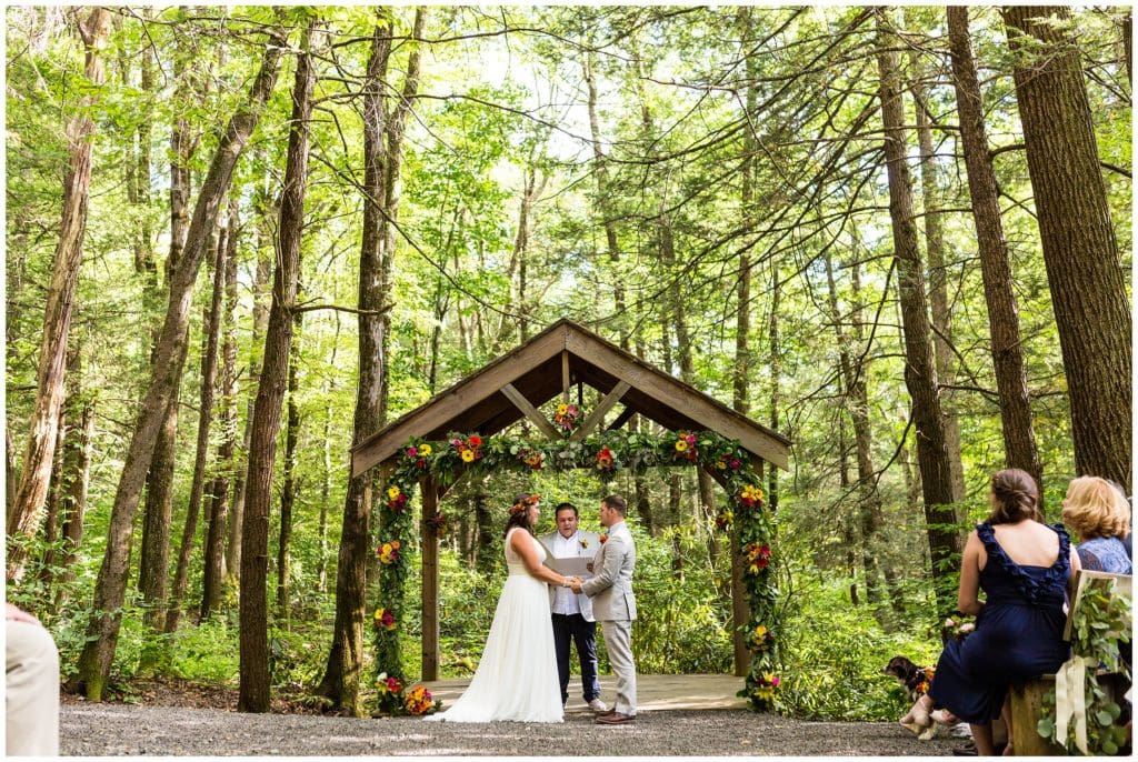 Outdoor woods wedding ceremony with bright floral arch and flower crowns, bride and groom at alter