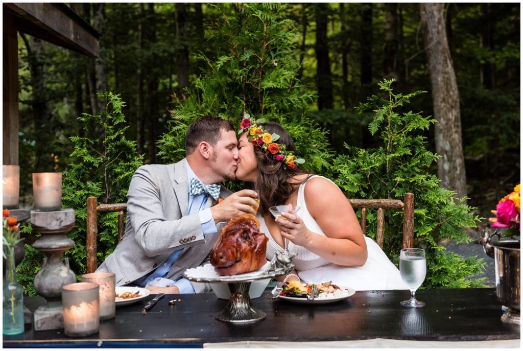 Bride and groom kissing and toasting at sweetheart table with pig roast head on table