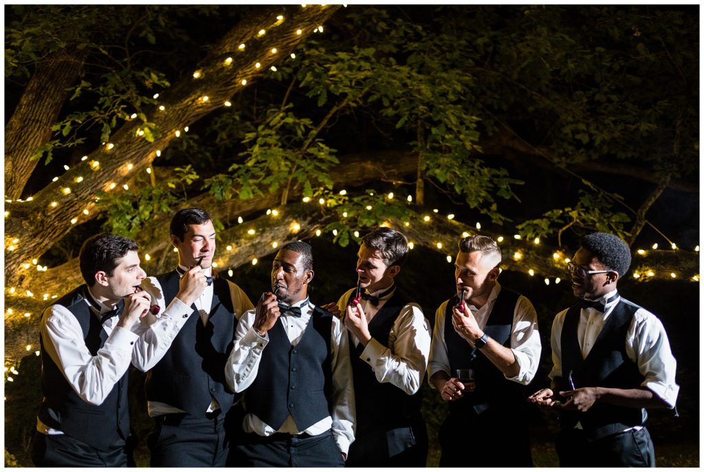 Groomsmen in vests and bowties smoke pipes with groom under light lined tree at Aldie Mansion wedding reception