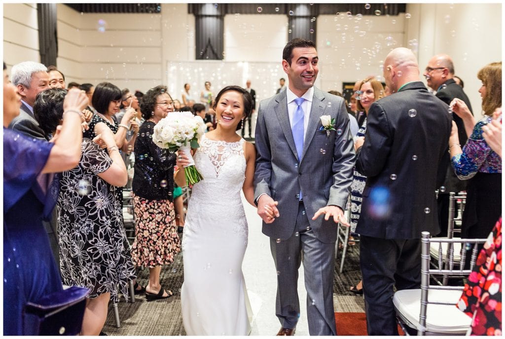 Bride and groom walk up aisle through guests blowing bubbles at Loews Philadelphia wedding ceremony