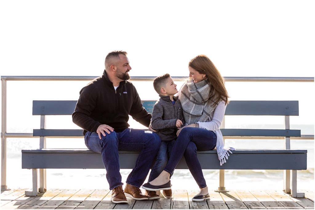 Couple sits on bench and laughs with son on Ocean City New Jersey Boardwalk engagement session