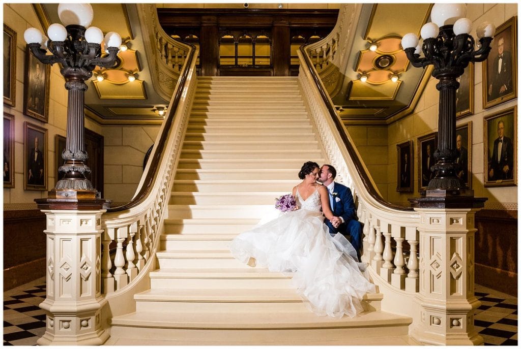 Bride and groom sit on grand stairway at One North Broad wedding reception