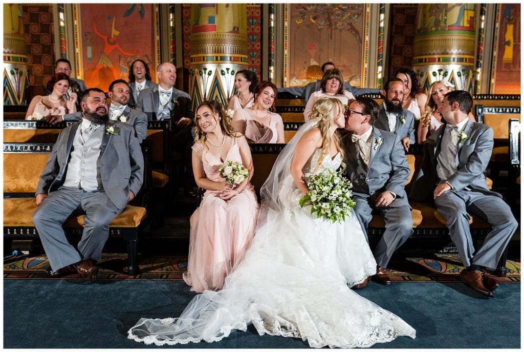 Wedding party laugh while bride and groom kiss in church pews portrait