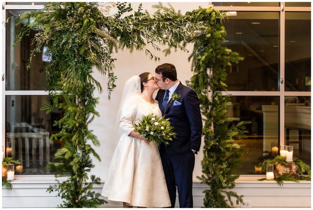 Bride and groom kiss at alter under pine and greenery arch at intimate Malvern Buttery wedding ceremony