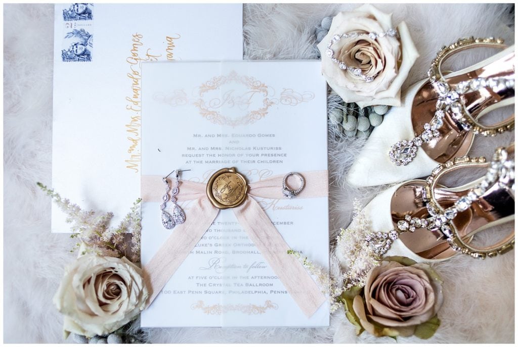 White pink and gold wedding invitation with gold wax seal and pink bowtie, white and gold wedding heels, earrings, engagement ring, and dusty pastel roses