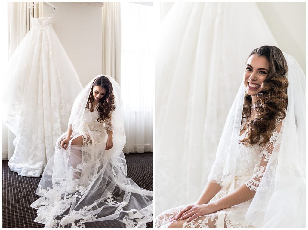 window lit bride in lace robe and long lace veil sitting in front of hanging wedding gown at Loews Philadelphia
