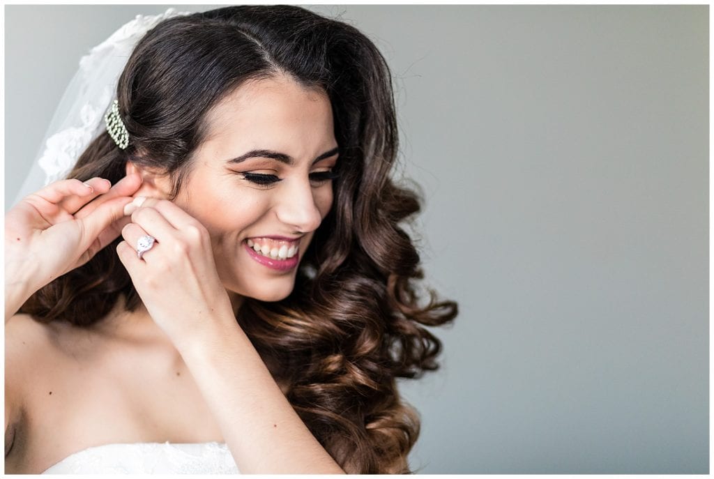 Window lit bridal portrait of bride putting on earrings