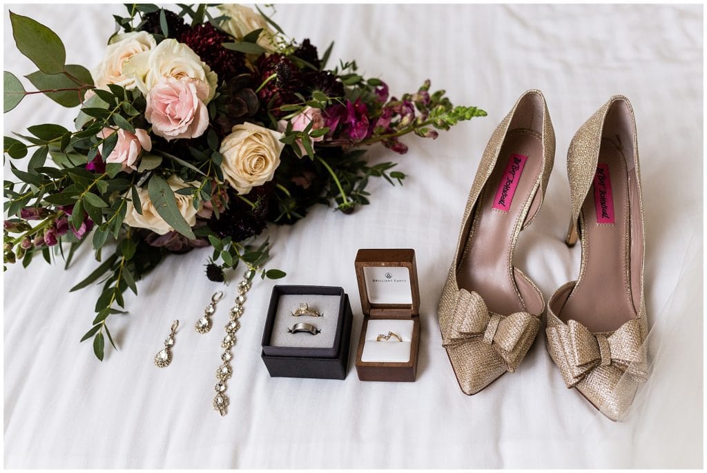 Bridal accessory details with gold bow heels, wedding bands and engagement ring, matching earrings and bracelet, and dark rose bouquet