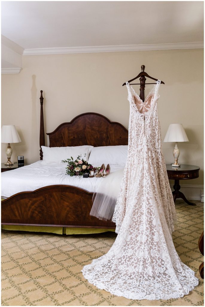 Lace wedding gown with long train hanging on bed post next to bouquet and bridal accessories