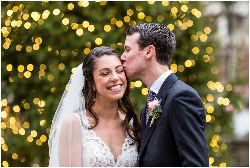 Groom kisses bride on cheek in front of tree with Christmas lights in Rittenhouse Square Park