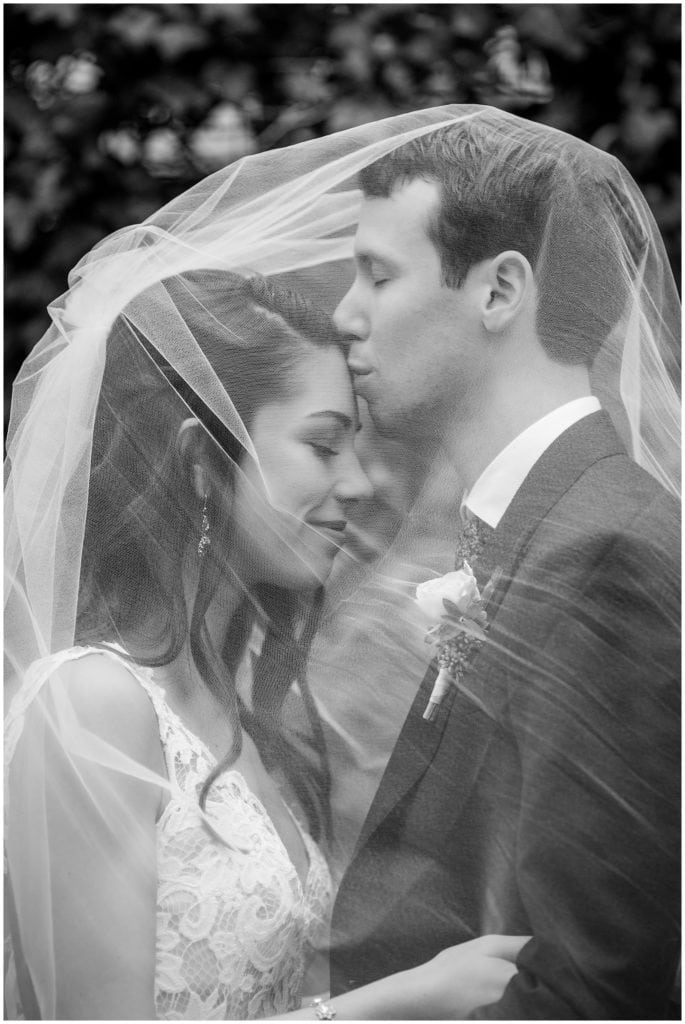 Groom kisses bride on forehead under veil black and white wedding portrait