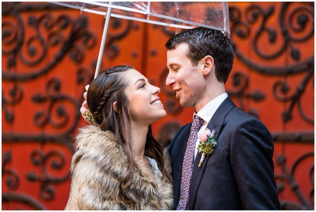 Bride and groom smile at each other under umbrella in front of old red doors in Center city Philadelphia winter holiday wedding