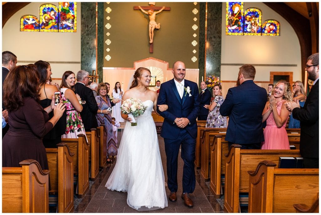 Bride and groom walk up aisle with guests cheering at St. Andrew's church wedding ceremony