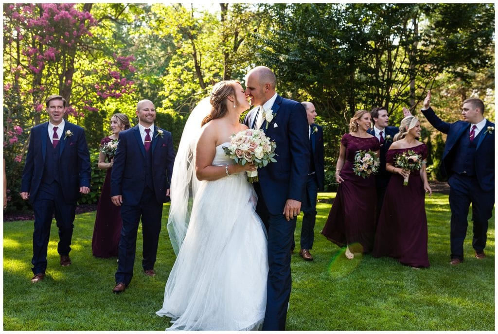 Bride and groom kiss while walking through garden with wedding party