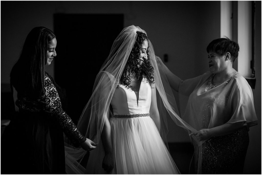 Black and white window lit portrait of mother of the bride and sister of the bride placing veil over brides head