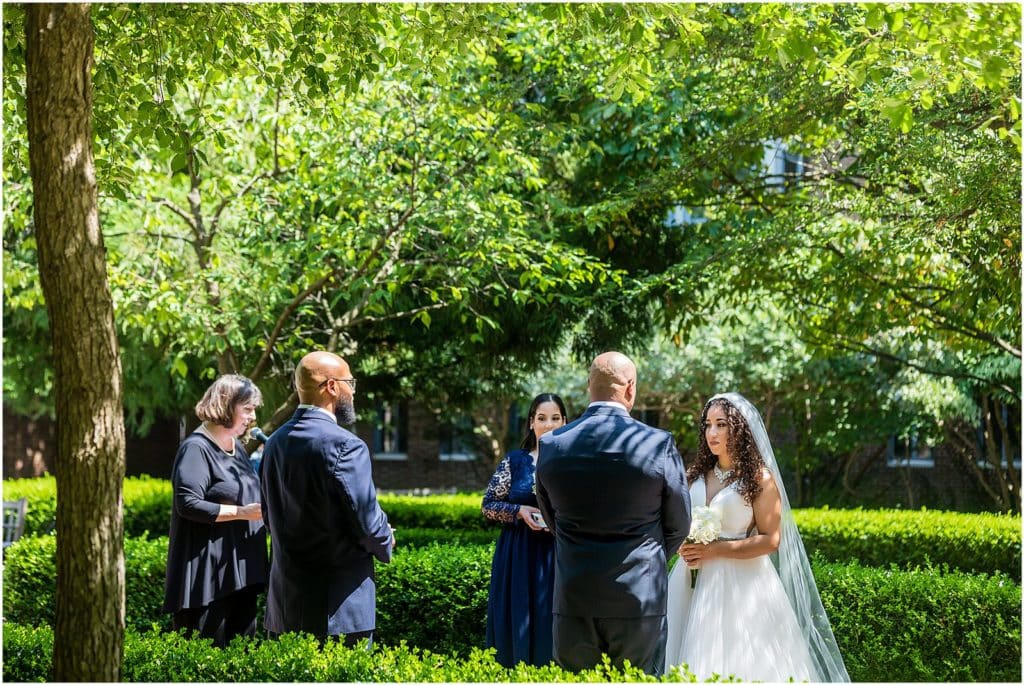 Bride and groom stand with maid of honor and best man during intimate outdoor ceremony in Penn Museum gardens micro wedding